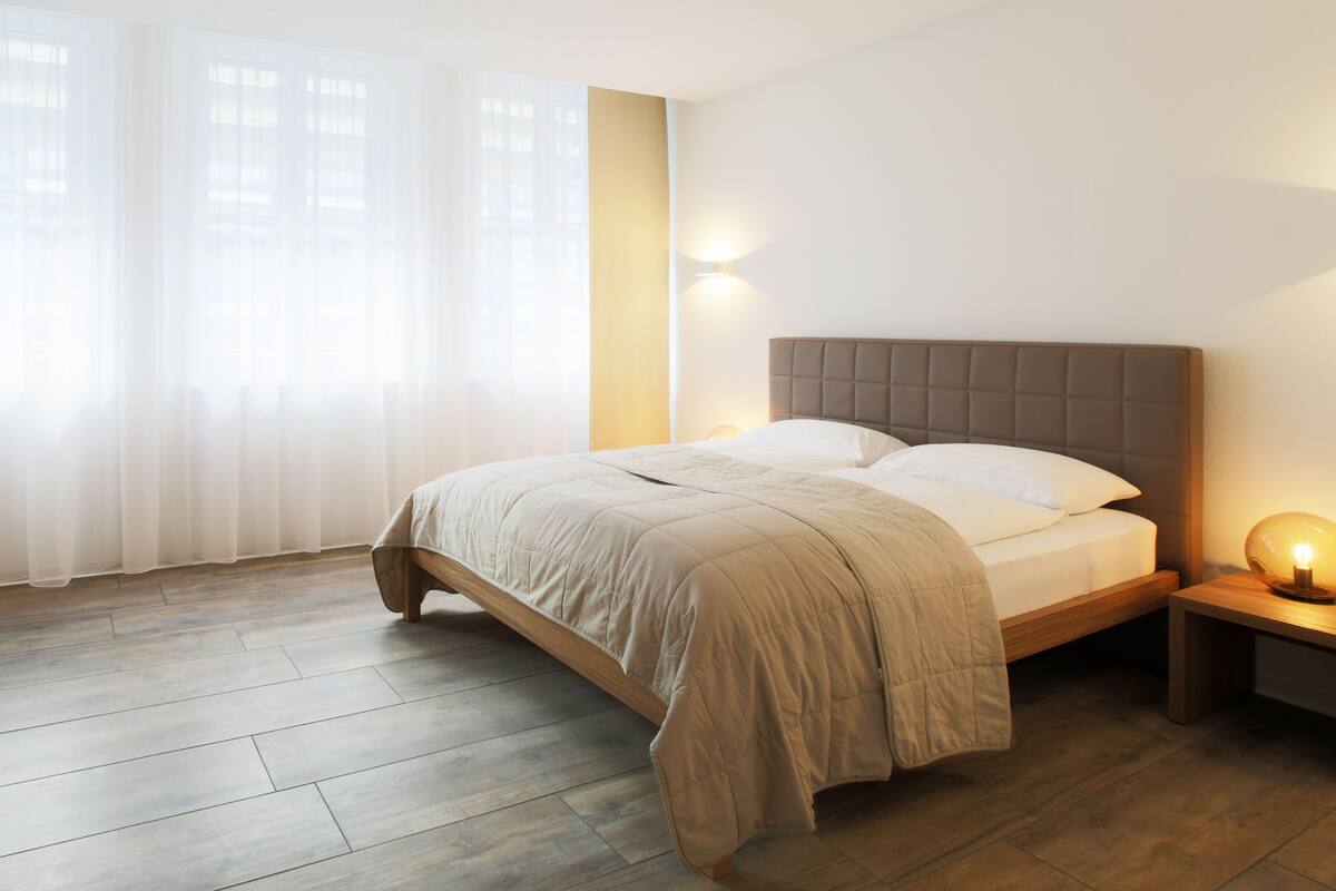 Un dormitorio con una cama, una mesita de noche con lámpara y una ventana con cortinas translúcidas.