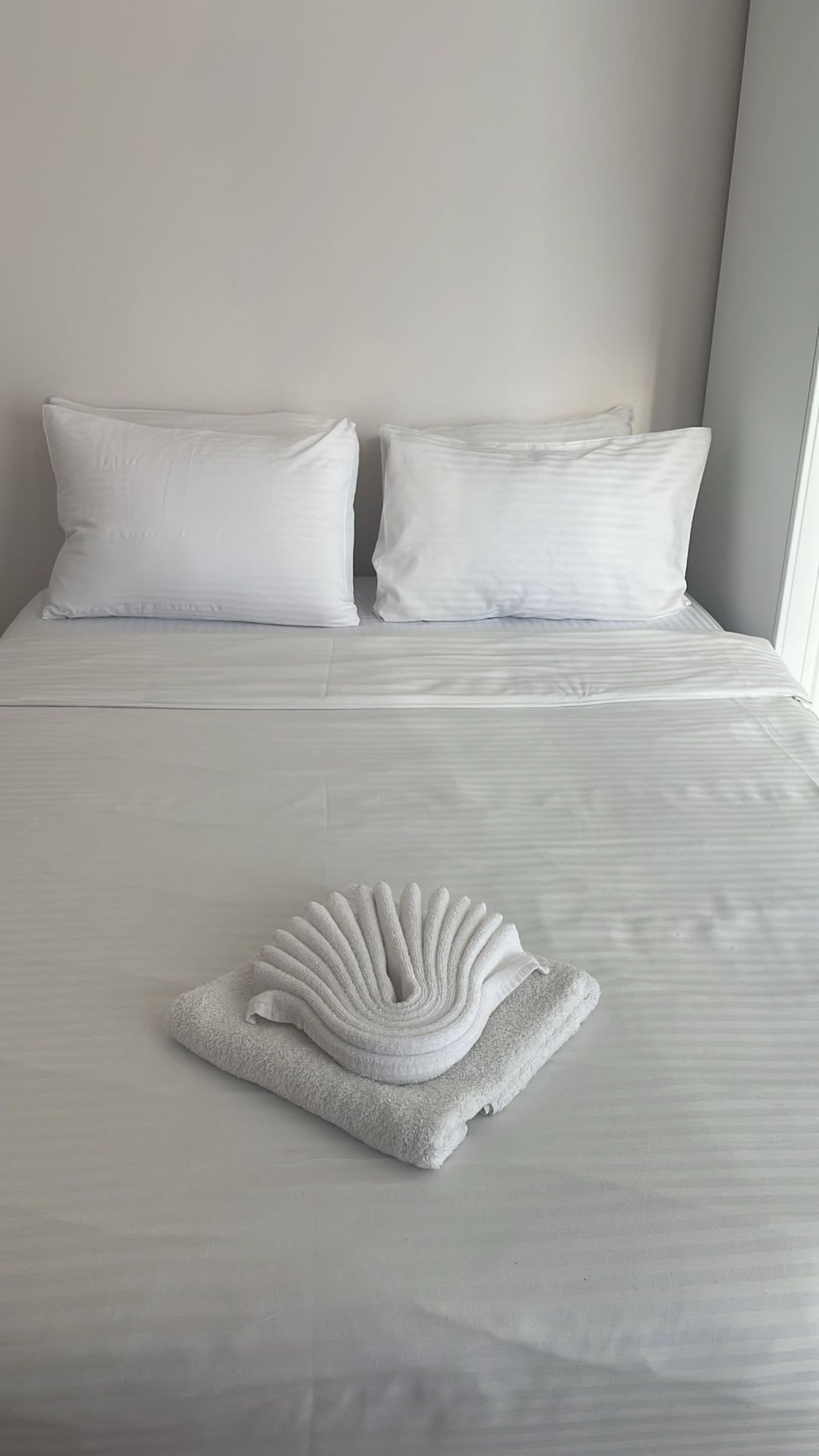 A neatly made bed with white linens and pillows, a folded towel, and a plain wall.