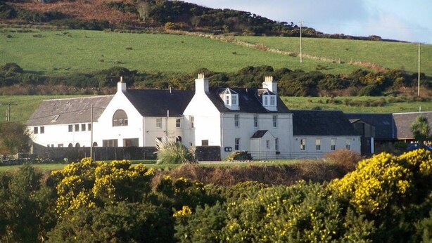 A large, white, two-story house with a symmetrical roofline and multiple chimneys, surrounded by green fields and a hill in the background. - Gigha Hotel (Isle of Gigha)