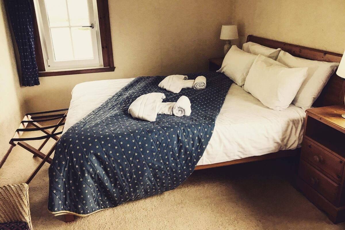 Ein ordentlich bezogenes Bett mit einer blauen Steudecke, zwei aufgerollten Handtüchern und einem Nachttisch mit einer Lampe.