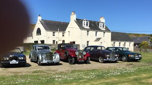 A row of vintage cars parked in front of a large, white building with multiple chimneys. - Gigha Hotel (Isle of Gigha)