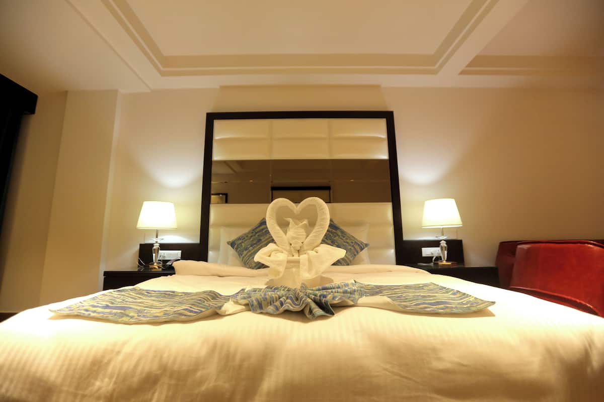 A hotel room with a large bed, two bedside lamps, a heart-shaped towel arrangement, and a mirror above the bed.