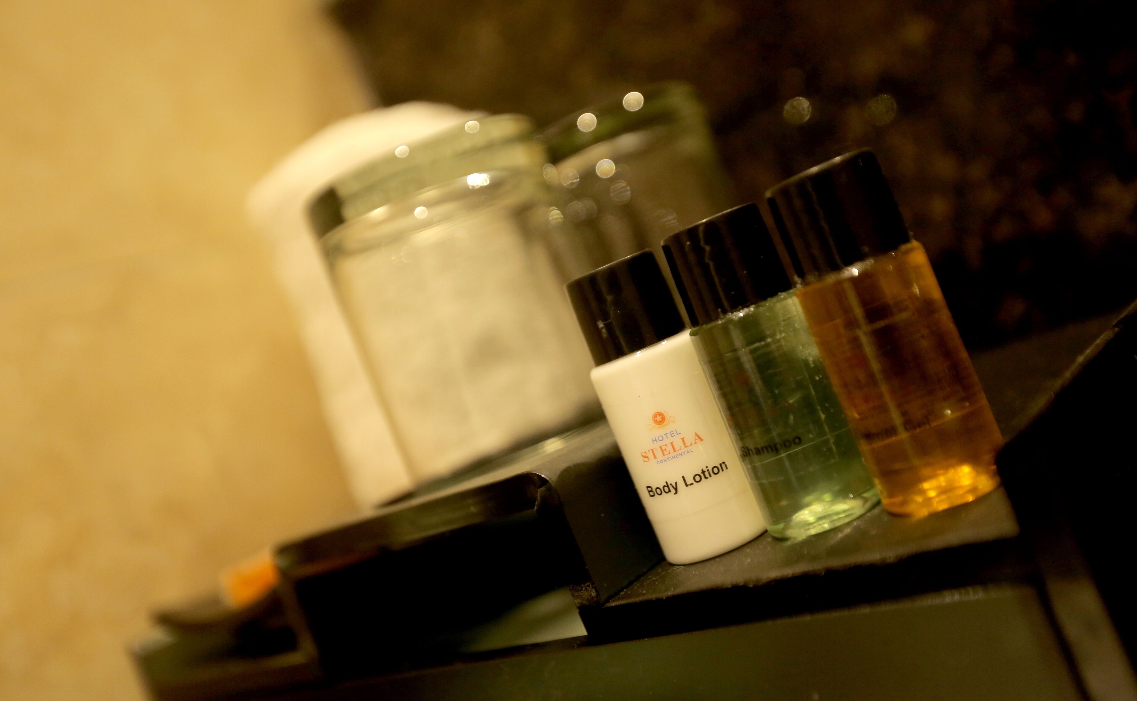 Una encimera de baño con artículos de aseo, incluyendo una botella etiquetada “Hotel Stella Continental Body Lotion”, un envase de champú y un tarro de crema hidratante.