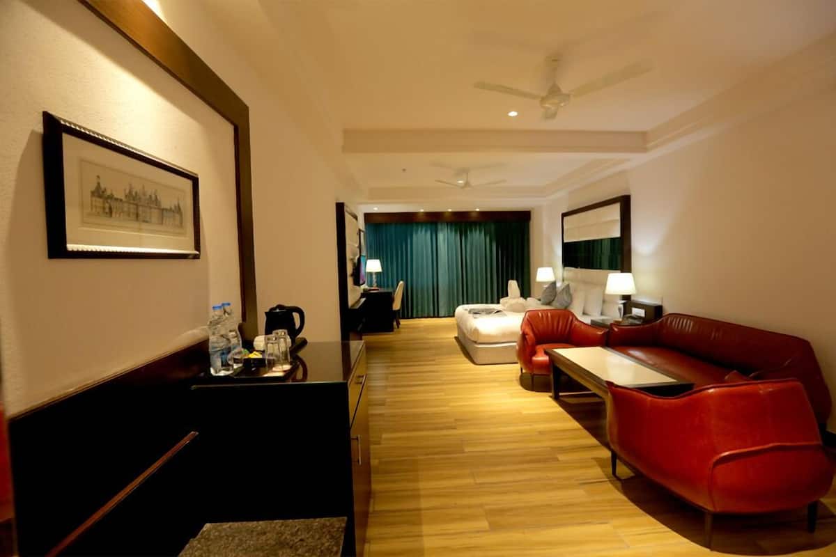 A hotel room with a wooden floor, a red leather sofa, a small table, a desk, a chair, a bed with a green curtain, and a ceiling fan.