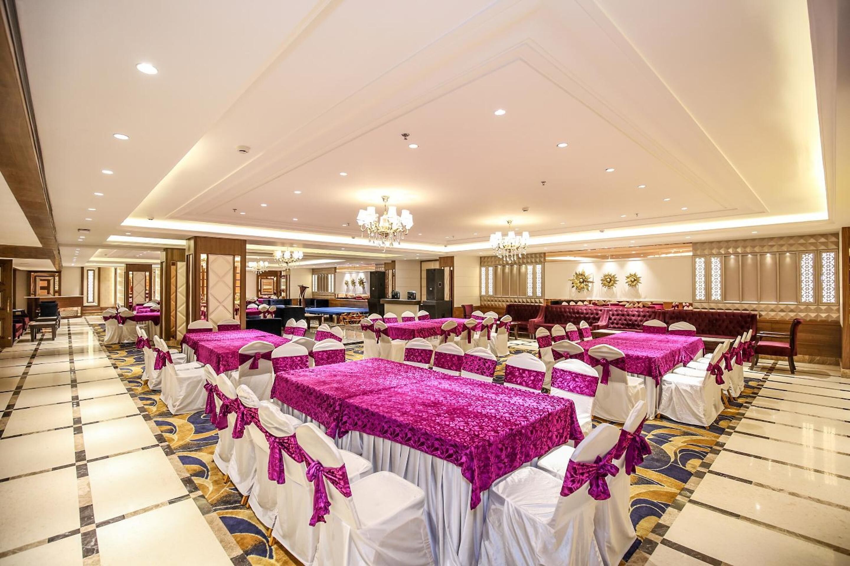 Une salle de banquet avec des rangées de tables recouvertes de nappes roses, des chaises blanches ornées de rubans roses et des lustres au plafond.