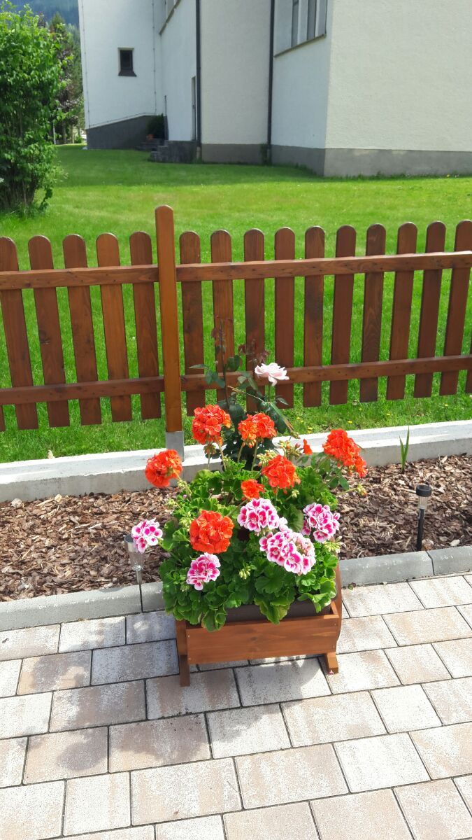A wooden planter box with flowers on a paved surface.