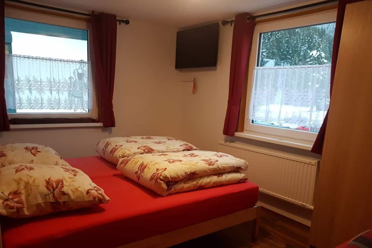 A bedroom with a bed, two windows with curtains, a mounted TV, and a radiator.