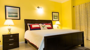 A neatly made bed with a white duvet and colorful pillows, a dark wooden headboard, and two bedside lamps on nightstands. - Kings Manor 06 by Pro Homes Jamaica (Kingston)