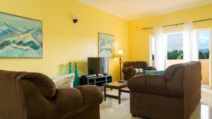 A living room with two brown sofas, a TV, and artwork on the walls. - Kings Manor 06 by Pro Homes Jamaica (Kingston)