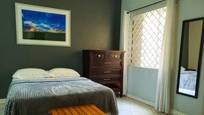 Ein Schlafzimmer mit einem Bett, einer Holzkommode, einem kleinen Holztisch, einem gemusterten Teppich, einem gerahmten Bild an der Wand und einem Fenster mit Vorhängen.