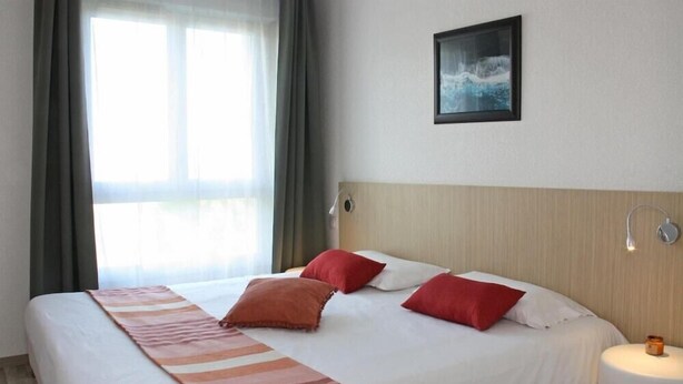 Una habitación de hotel con cama, ventana con cortinas, un cuadro en la pared y una mesita de noche con lámpara.