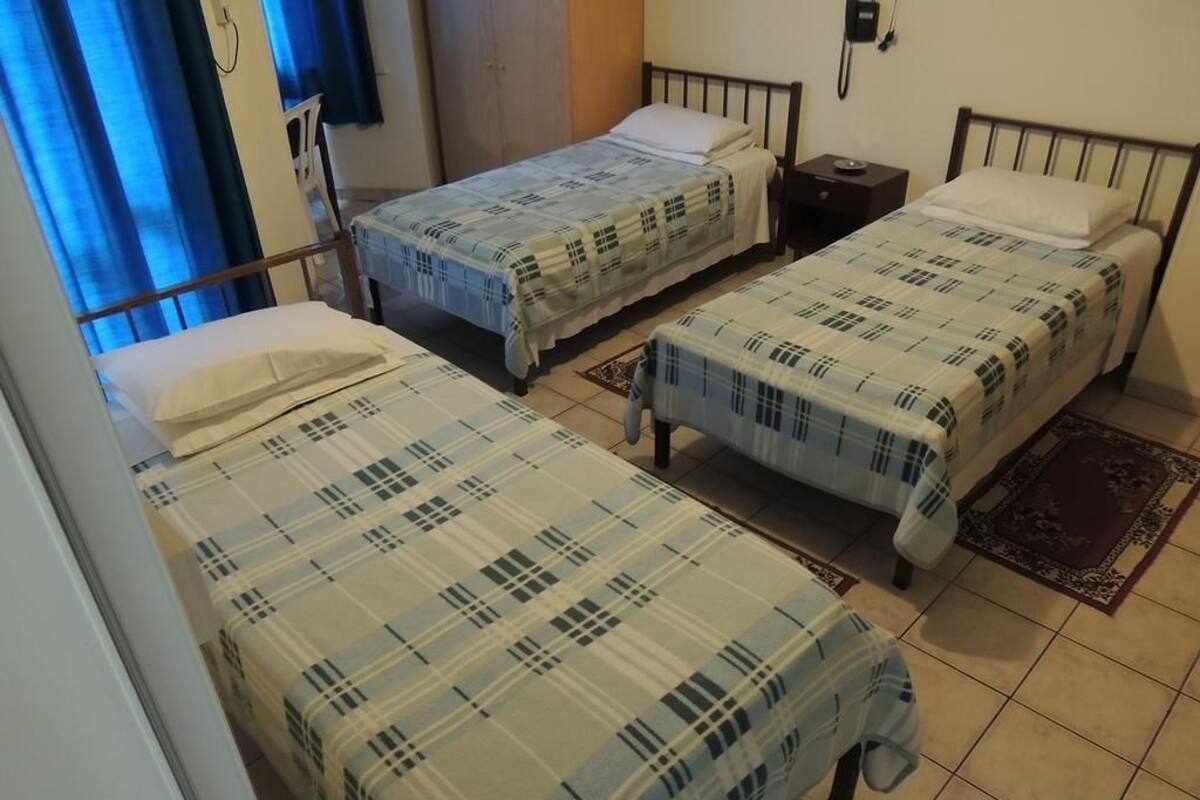 Une chambre avec trois lits simples, chacun étant équipé d’une couette et d’un oreiller assortis. On y trouve une petite table de chevet avec une lampe d’un côté et un téléphone de l’autre. La chambre a un sol carrelé et une fenêtre avec des rideaux bleus.