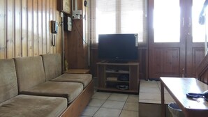 A wooden-paneled room with a sofa, a television on a wooden stand, a table, and a window with blinds. - Delphi Hotel (Nicosia)