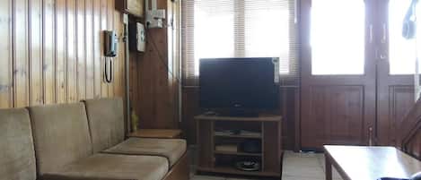 A wooden-paneled room with a sofa, a television on a wooden stand, a table, and a window with blinds.