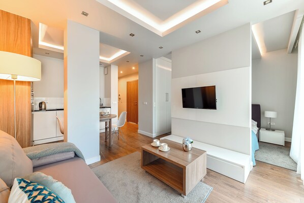 A modern living room with a sofa, coffee table, and TV. There is a kitchen area in the background with a dining table and chairs. - P&O Oxygen Wronia 3 (Warsaw)