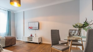 A modern living room with a sofa, a dining table with chairs, a TV mounted on the wall, and a potted plant. - P&O Chimielna 3 (Warsaw)