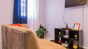 Una sala de estar con un sofá, un televisor de pantalla plana montado en la pared, una estantería con adornos y una planta en maceta.