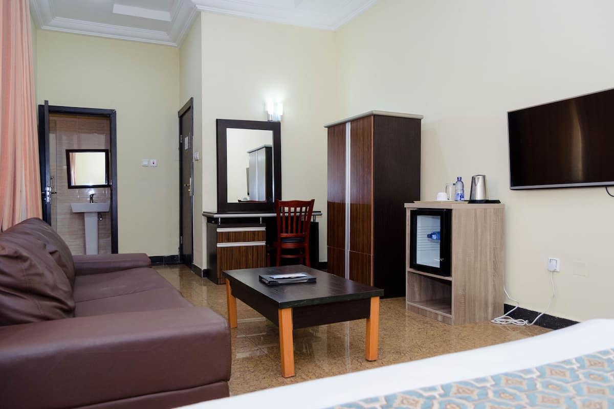 Een hotelkamer met een bruine leren bank, een houten bijzettafeltje, een bureau met stoel, een kledingkast, een televisie en een klein kastje met waterkoker en waterflessen.