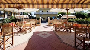 An outdoor dining area with tables and chairs under striped canopies, a tiled floor, and a building in the background. - Hotel Losanna (Cervia)