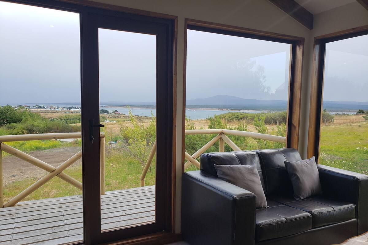 A room with a black leather sofa and a wooden coffee table, large windows with a view of a beach and mountains.
