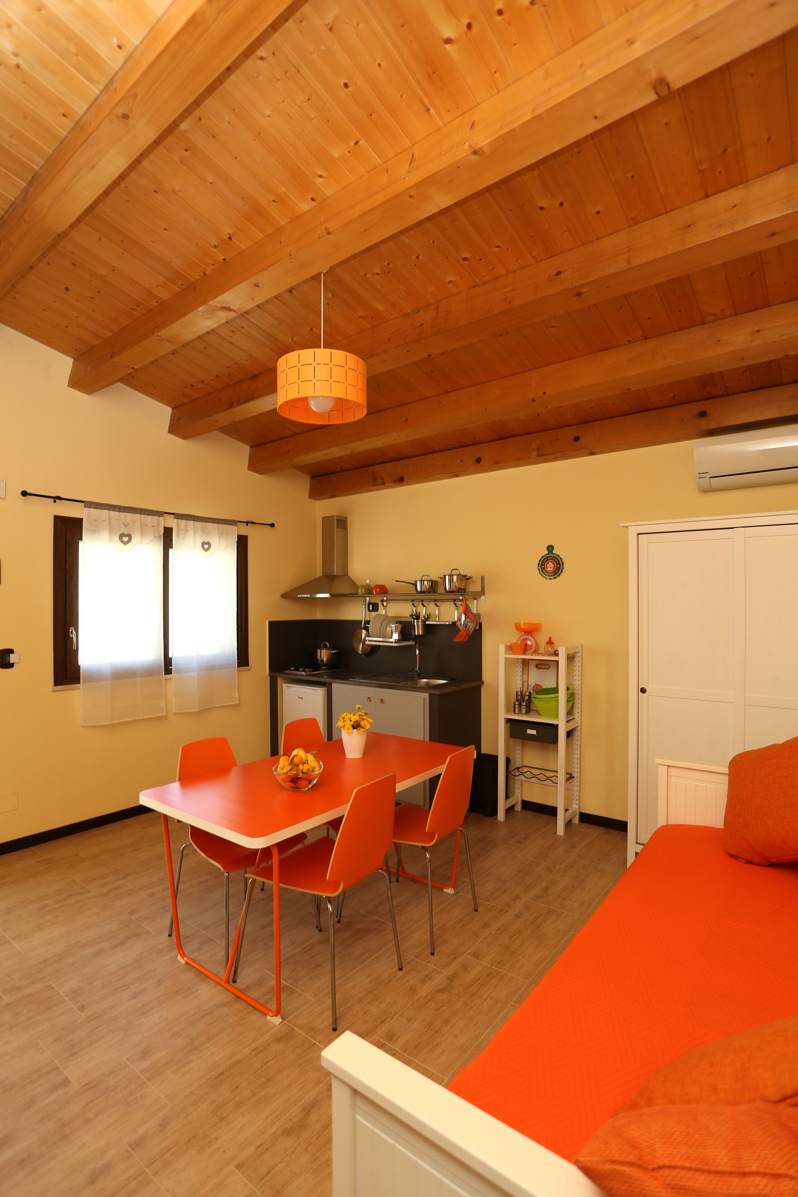 Habitació compacta amb sostre de fusta, taula de dinar amb cadires taronja, llit amb roba de llit taronja i una cuineta.