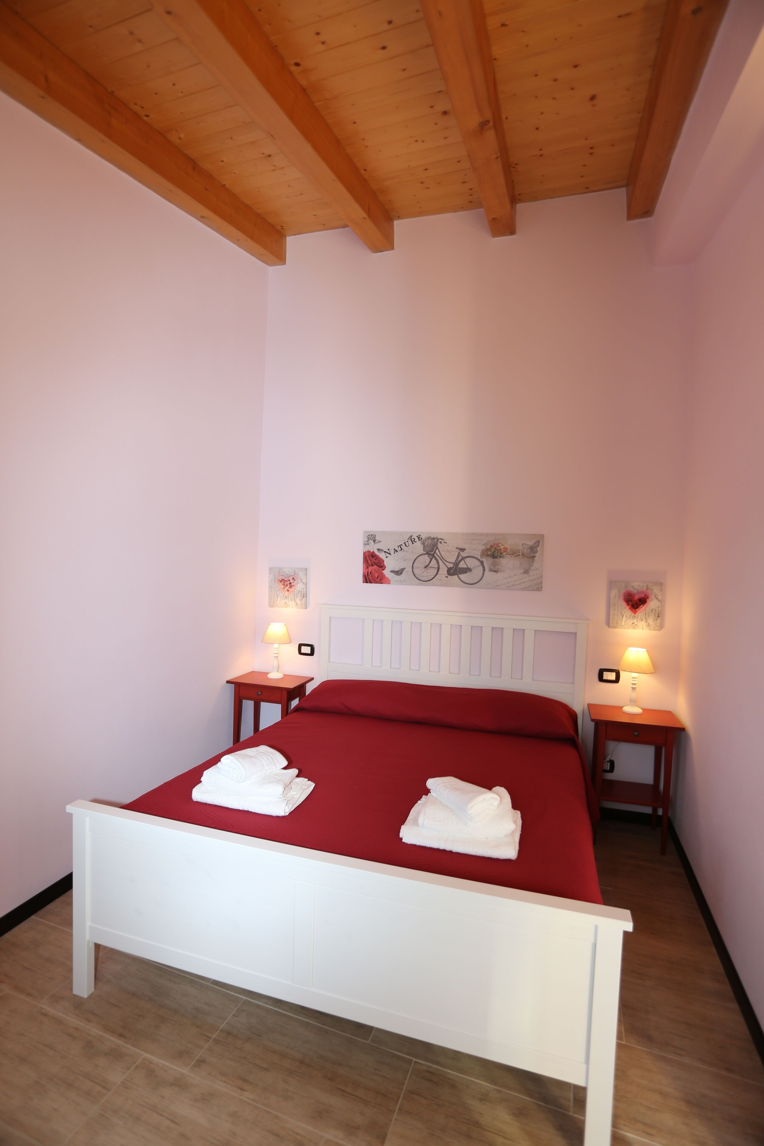 A bedroom with a white bed, red bedding, wooden ceiling beams, and two bedside tables with lamps.