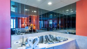 A modern bathroom with a large mirror, a jacuzzi tub, and a visible seating area in the background. - Fullerton Manor Inn (Niagara Falls)