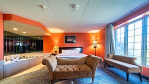 A hotel room with a large bed, two armchairs, a sofa, a large window, a wall-mounted TV, and a bathroom with a bathtub and a mirror. - Fullerton Manor Inn (Niagara Falls)