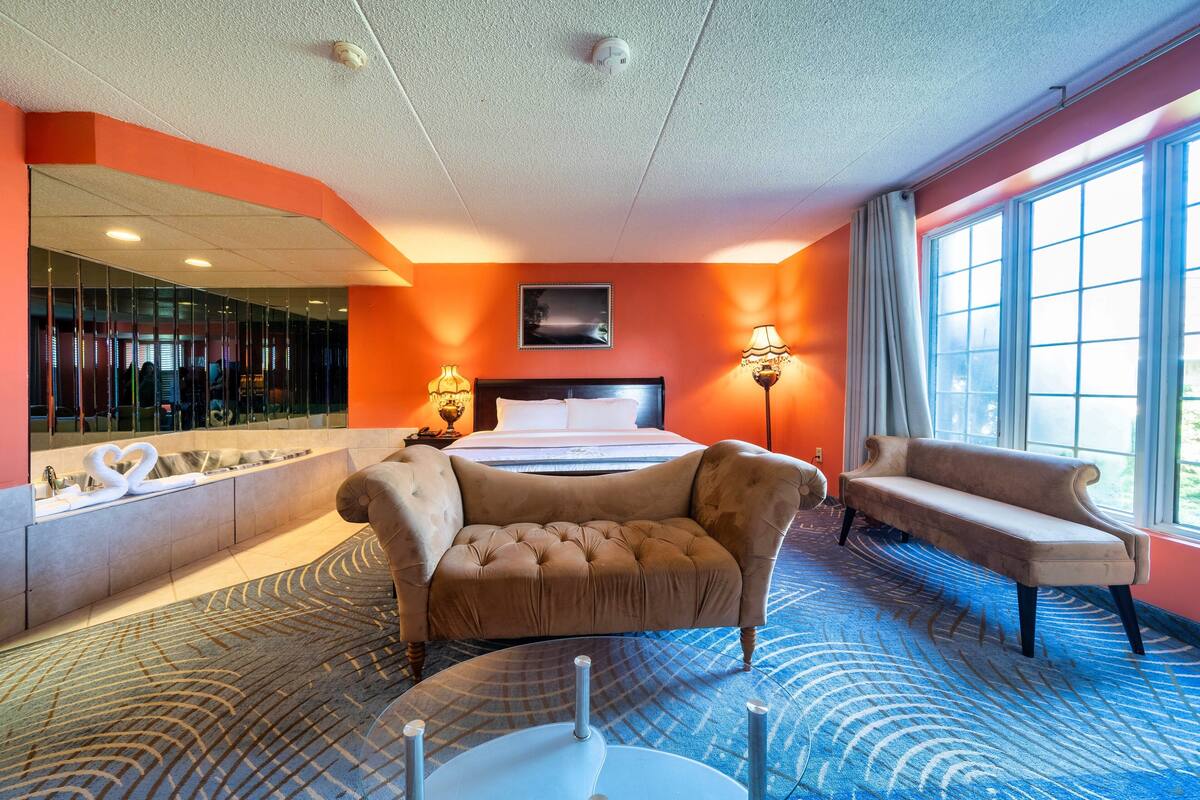 A hotel room with a large bed, two armchairs, a sofa, a large window, a wall-mounted TV, and a bathroom with a bathtub and a mirror.