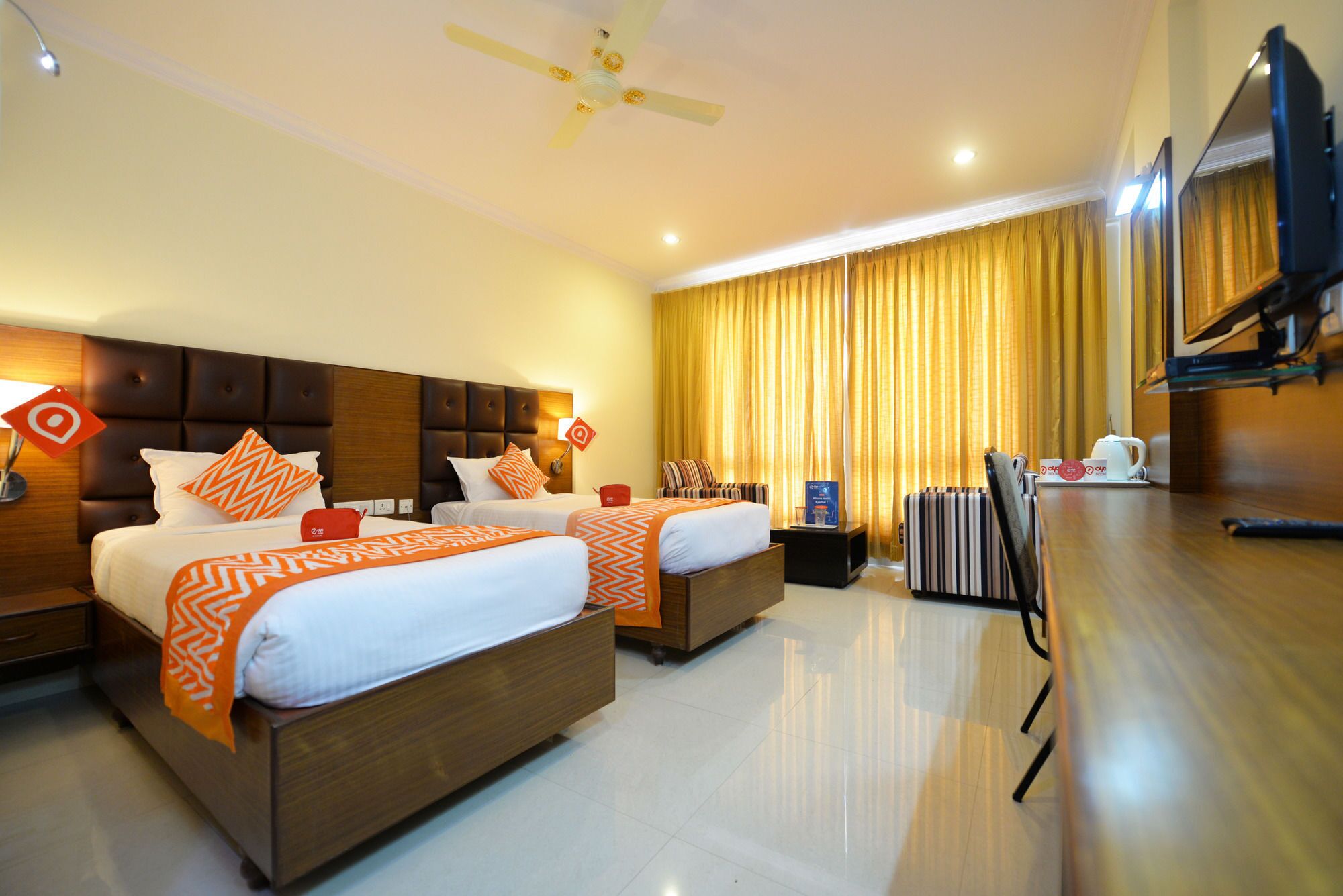 A hotel room with two beds, a flat-screen TV, a ceiling fan, and a large window with curtains.