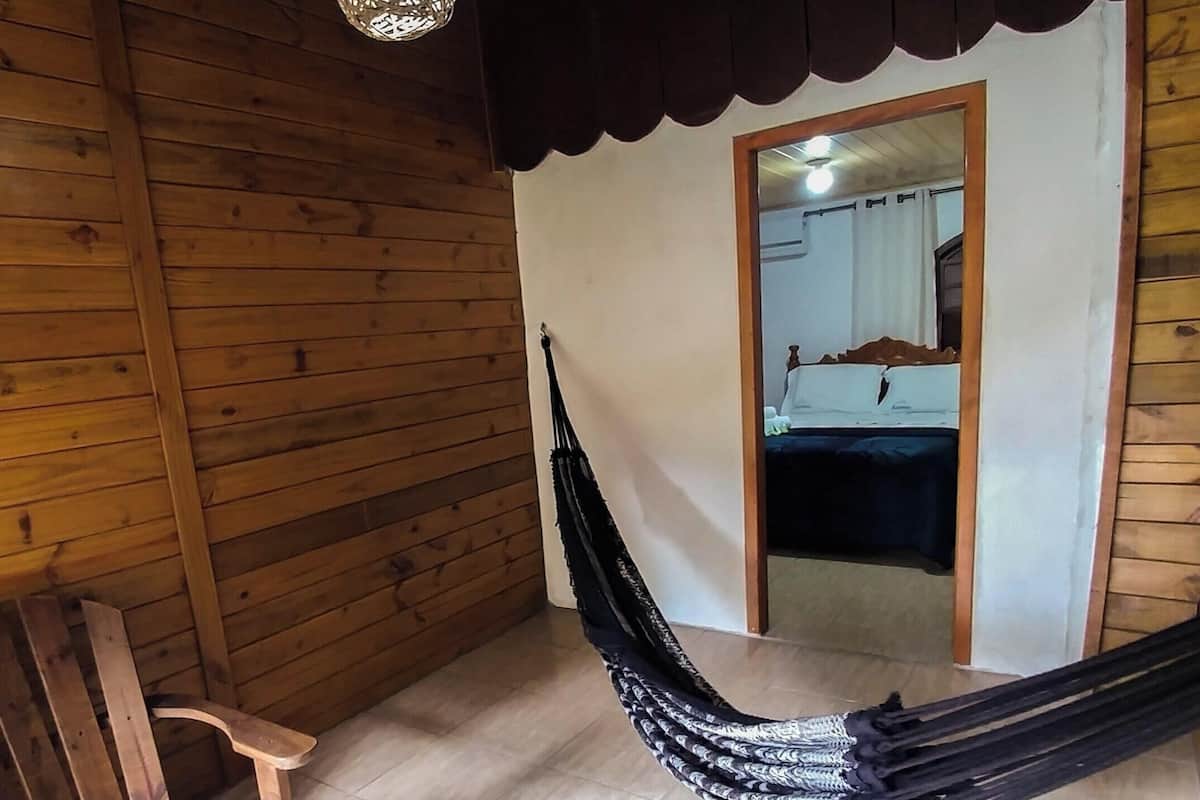 Interior em madeira com uma rede, um banco de madeira e uma porta que dá para outro quarto.
