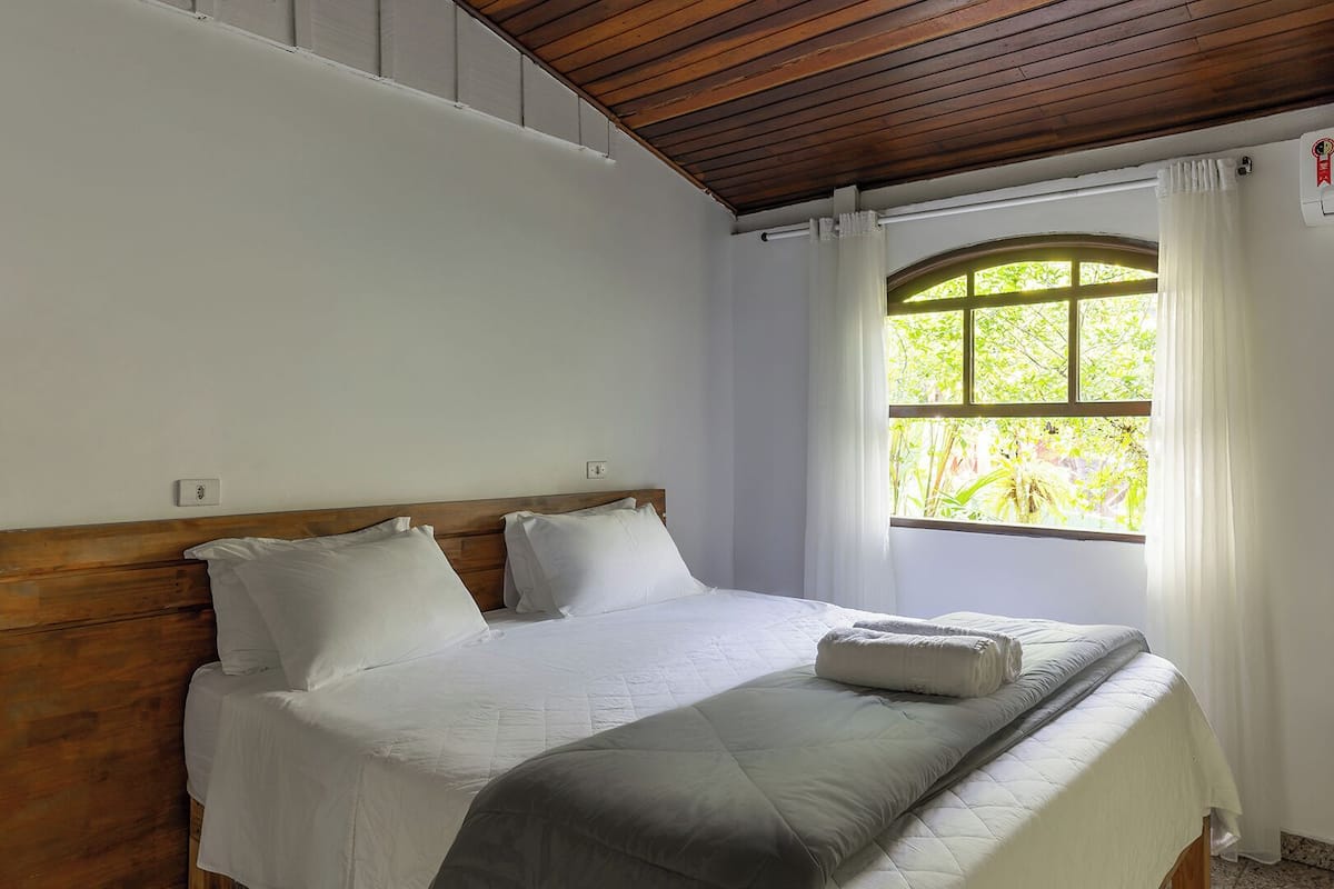 Quarto com cabeceira de madeira, uma janela grande com cortinas e cama com roupa de cama branca.