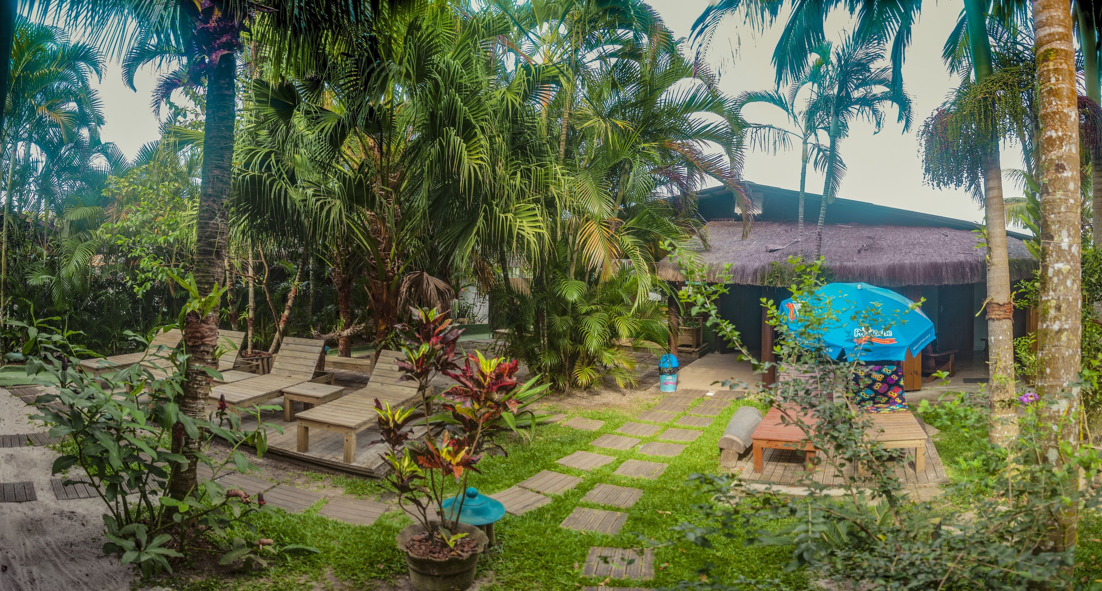 Un jardín tropical con una cabaña de techo de paja, terraza de madera y plantas en macetas.