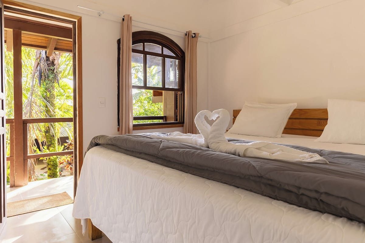 Quarto com cama, janela com cortinas e vista para o verde lá fora.