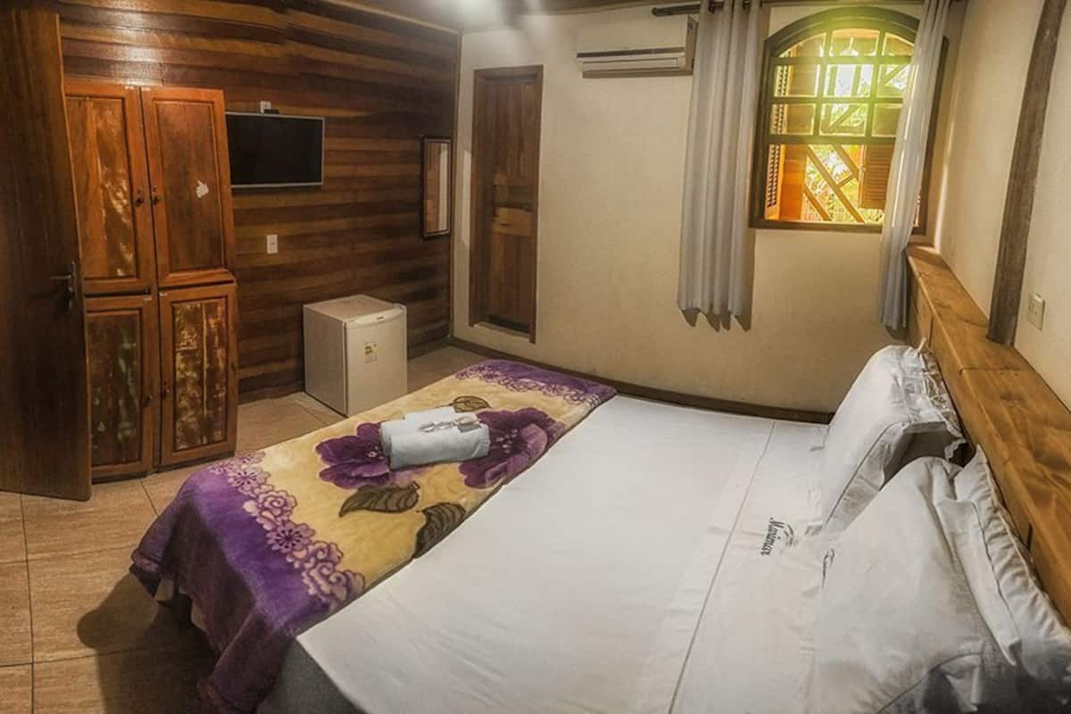 Quarto com cama, uma pequena escrivaninha, televisão e janela com cortinas.