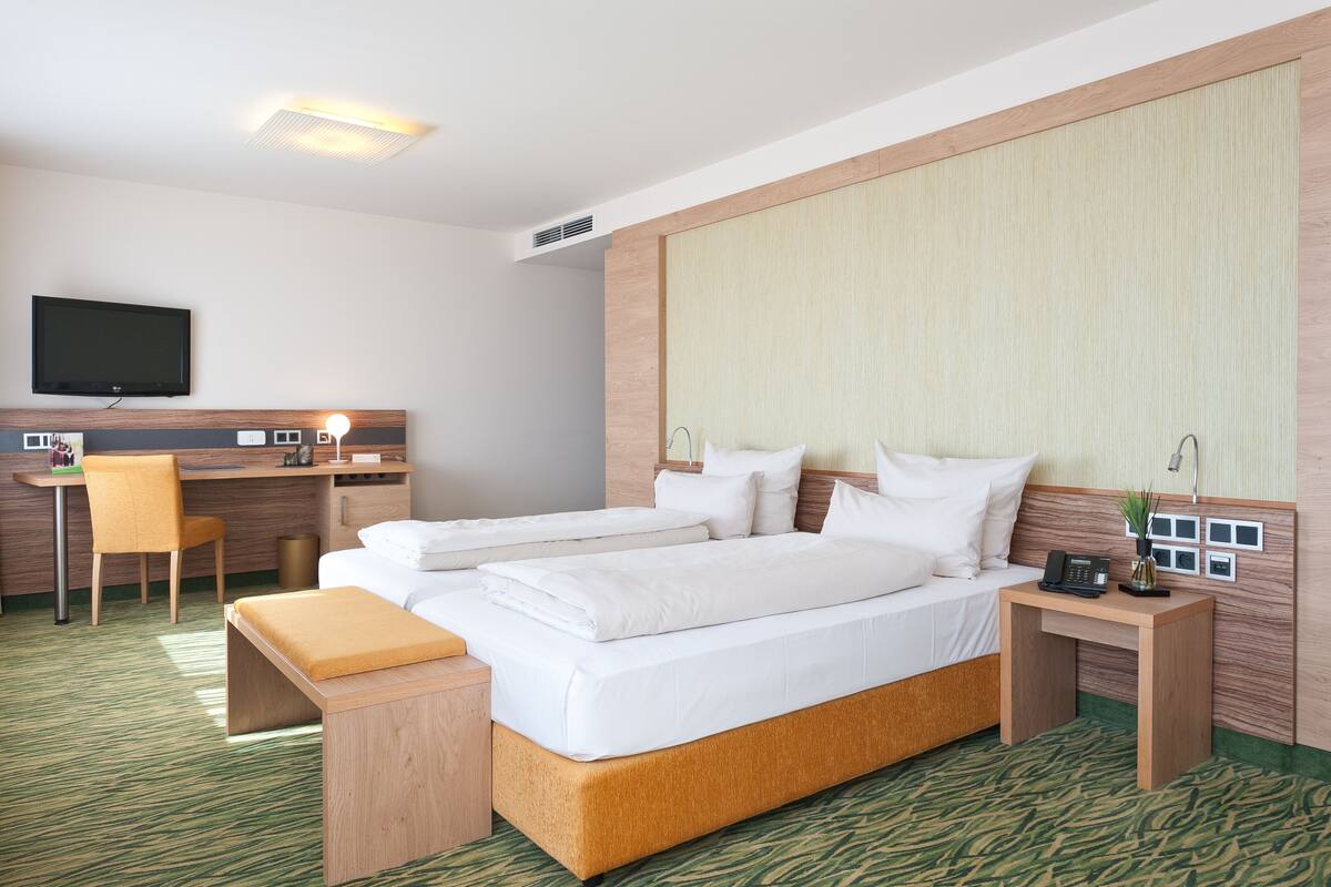 A hotel room with a bed, a desk, a chair, a TV, and a bedside table with a lamp.