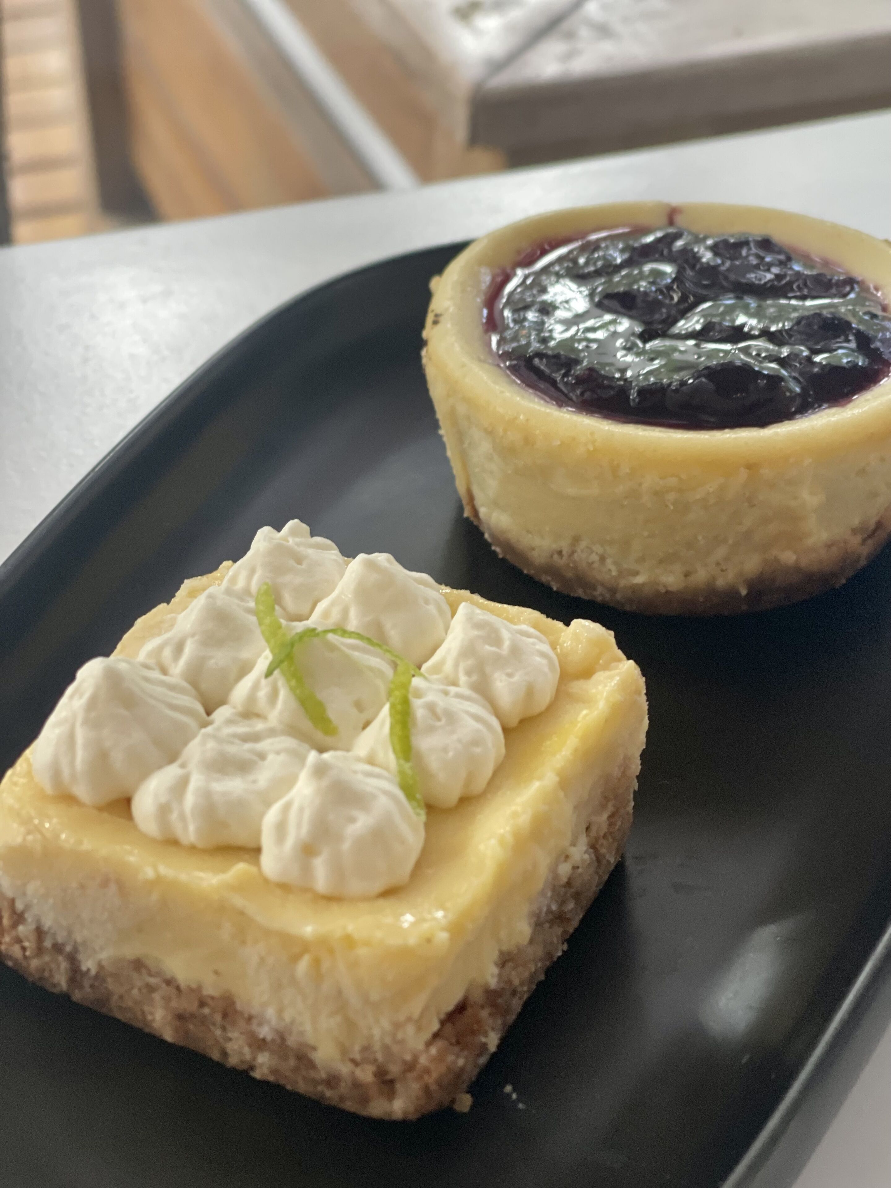 A dessert on a black plate, consisting of a cheesecake with whipped cream and a berry compote.