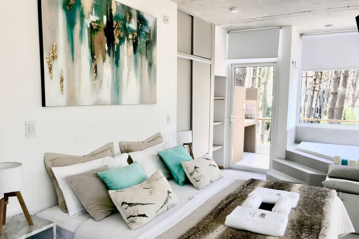 A modern bedroom with a large bed, a painting, and a view of the outdoors.