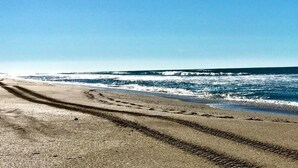 Una playa con huellas que conducen al agua, cielo despejado y olas rompiendo en la orilla.
