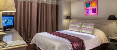 A hotel room with a bed, a desk with a lamp, a television, and a wall with colorful artwork.