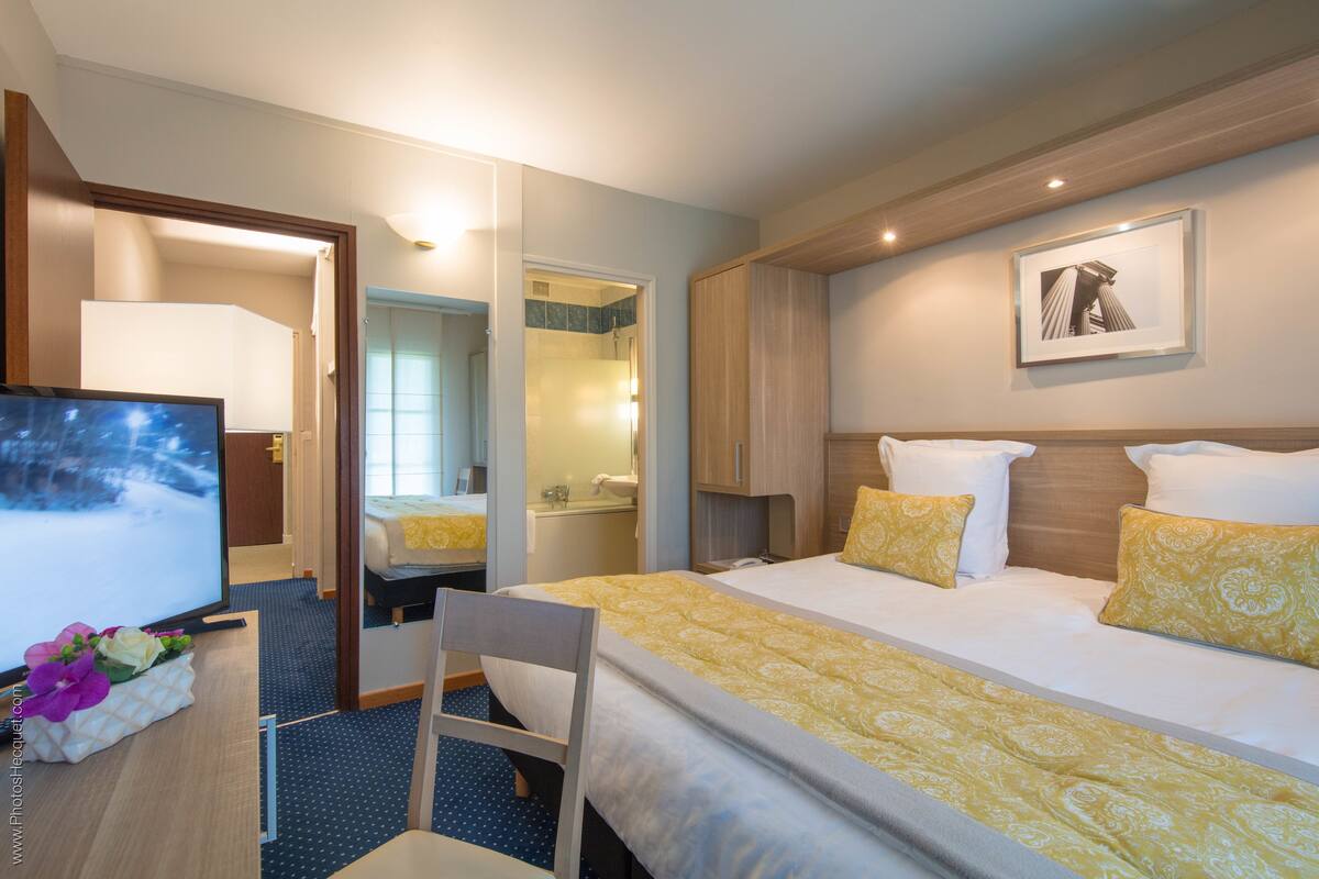 Ein Hotelzimmer mit einem großen Bett, einem Fernseher, einem Schreibtisch mit Blumen und einem durchgegangenen Tür sichtbaren Badezimmer.