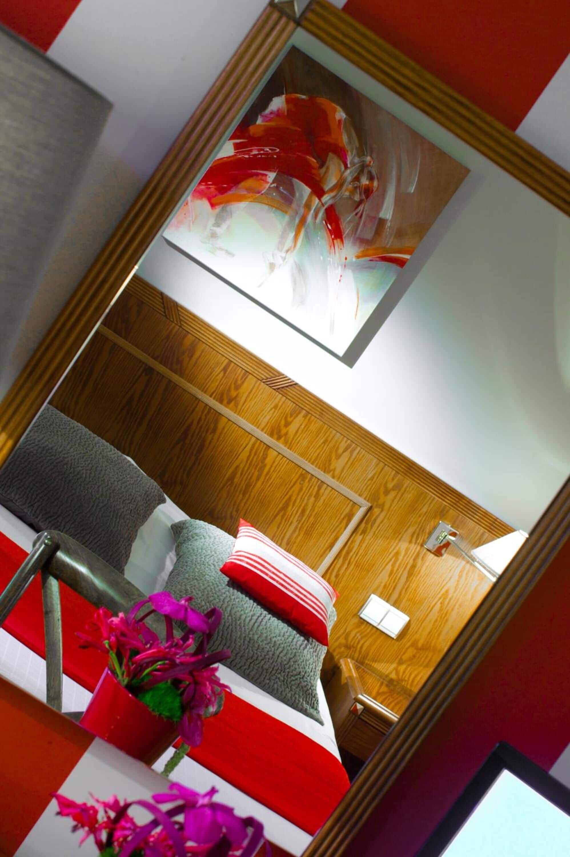 A modern bedroom with a wooden headboard, a bed with grey and red bedding, a bedside table with a lamp, and a mirror reflecting the room.