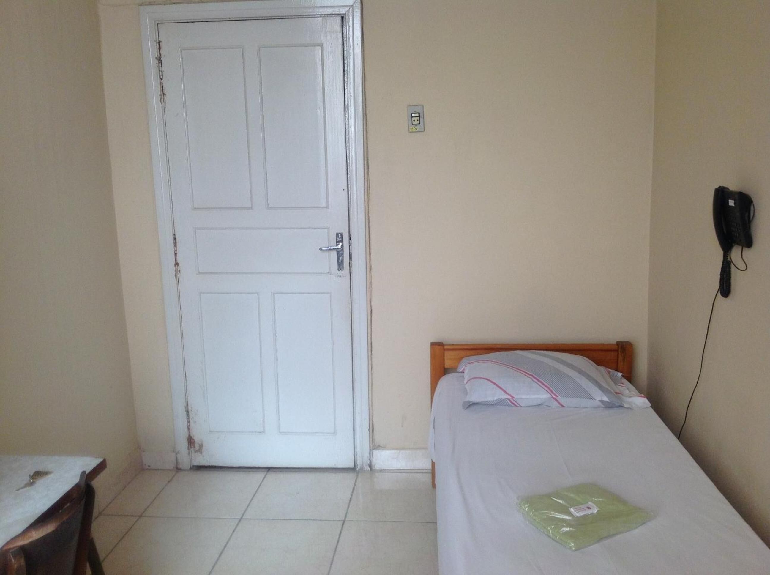 Una habitación pequeña y sencilla con una cama individual, una puerta, un teléfono en la pared y suelo de baldosas.