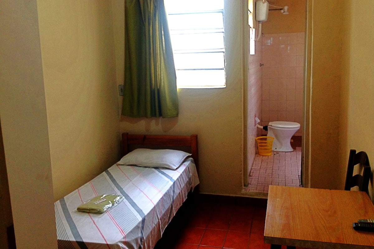 Quarto pequeno com cama, mesa, janela com cortinas e sanitário.