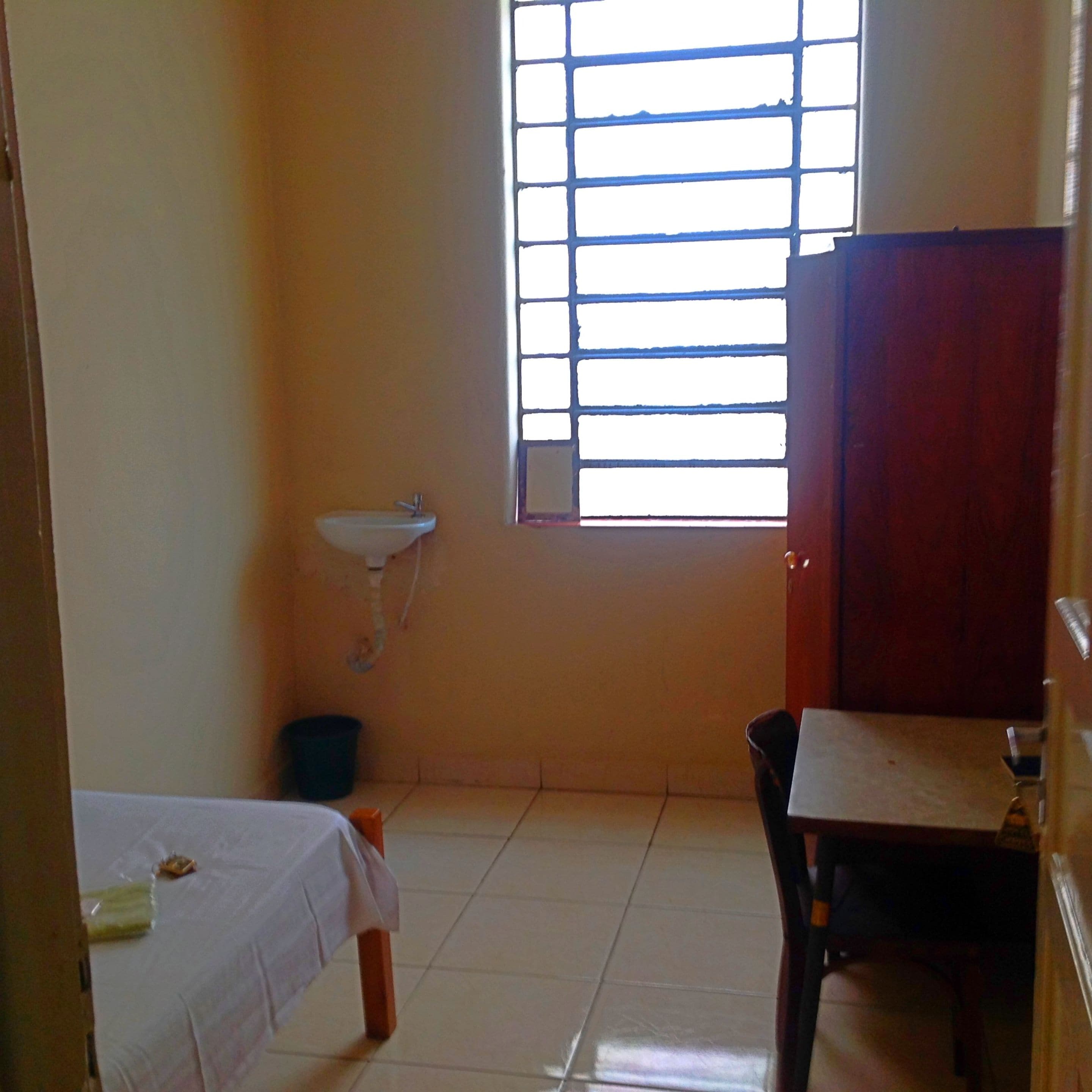 A small room with a bed, a table, a window with blinds, a sink, and a trash bin.