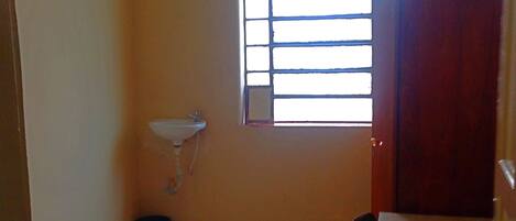 A small room with a bed, a table, a window with blinds, a sink, and a trash bin.