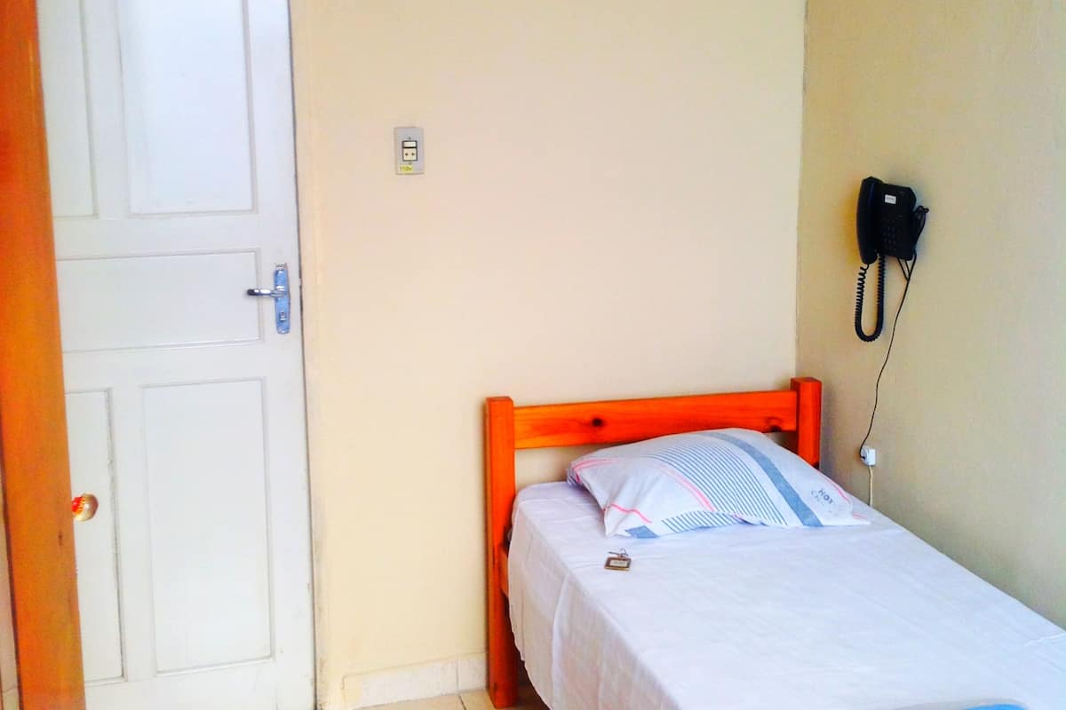 Quarto pequeno com uma cama de solteiro, cabeceira de madeira, porta branca, telefone fixado na parede e piso de azulejos.