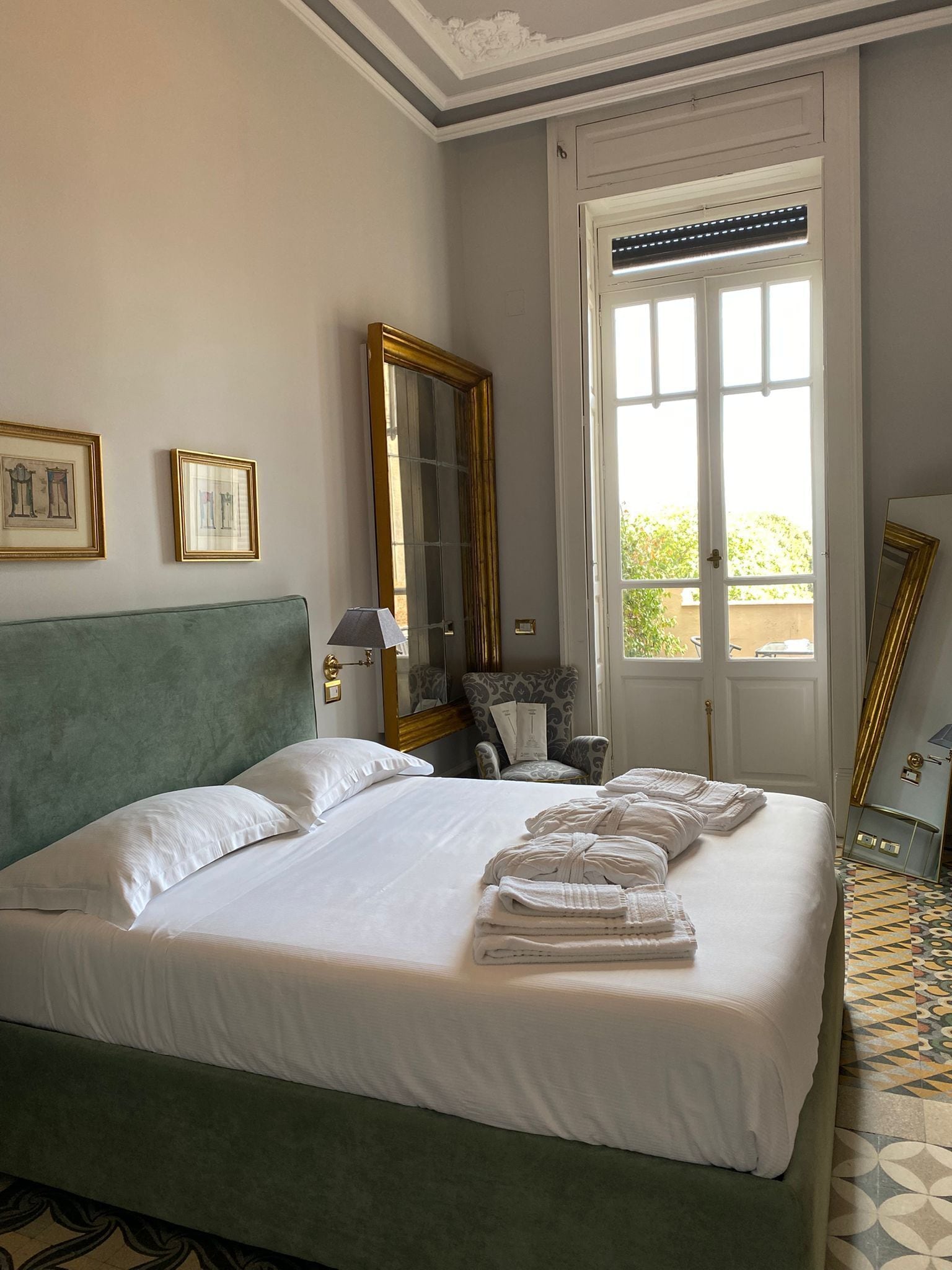 A neatly made bed with white linens and towels, a green upholstered headboard, a large mirror, and a window with a view of greenery outside.