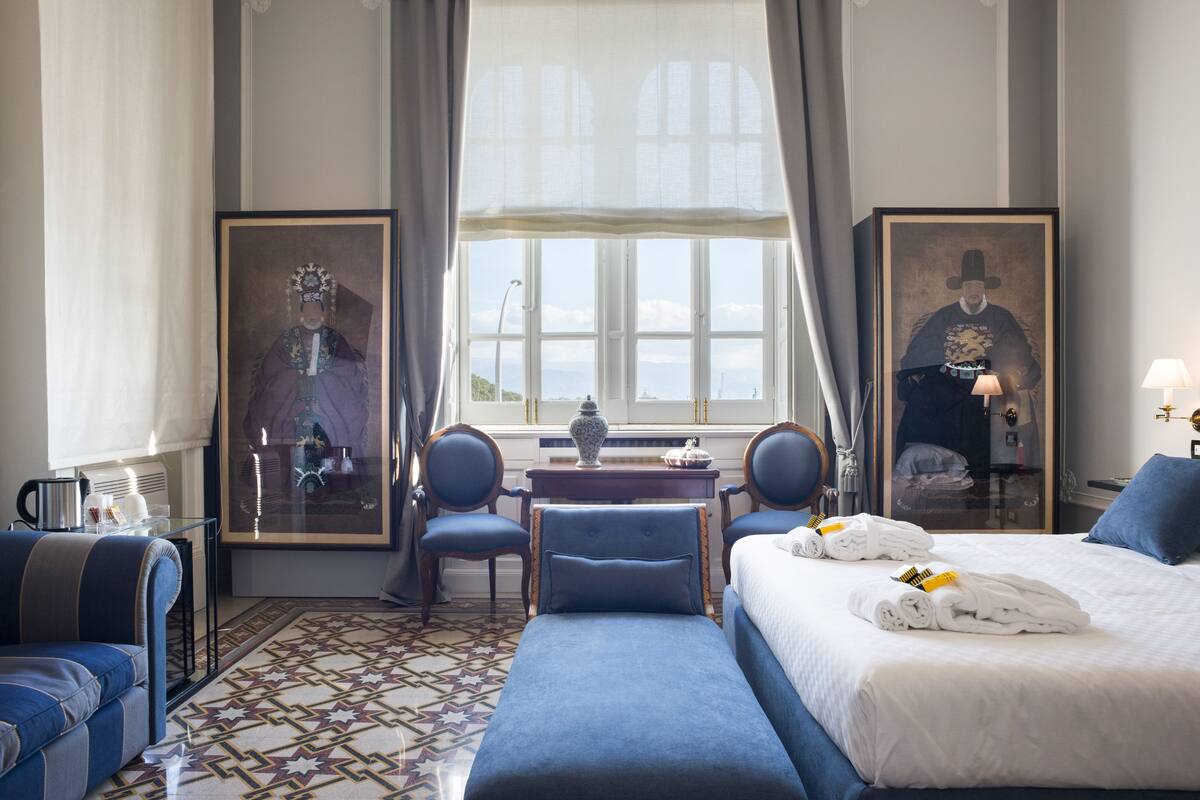 Une chambre d’hôtel avec un grand lit, un canapé, un bureau agrémenté d’un vase, deux chaises et une fenêtre donnant sur un beau paysage.