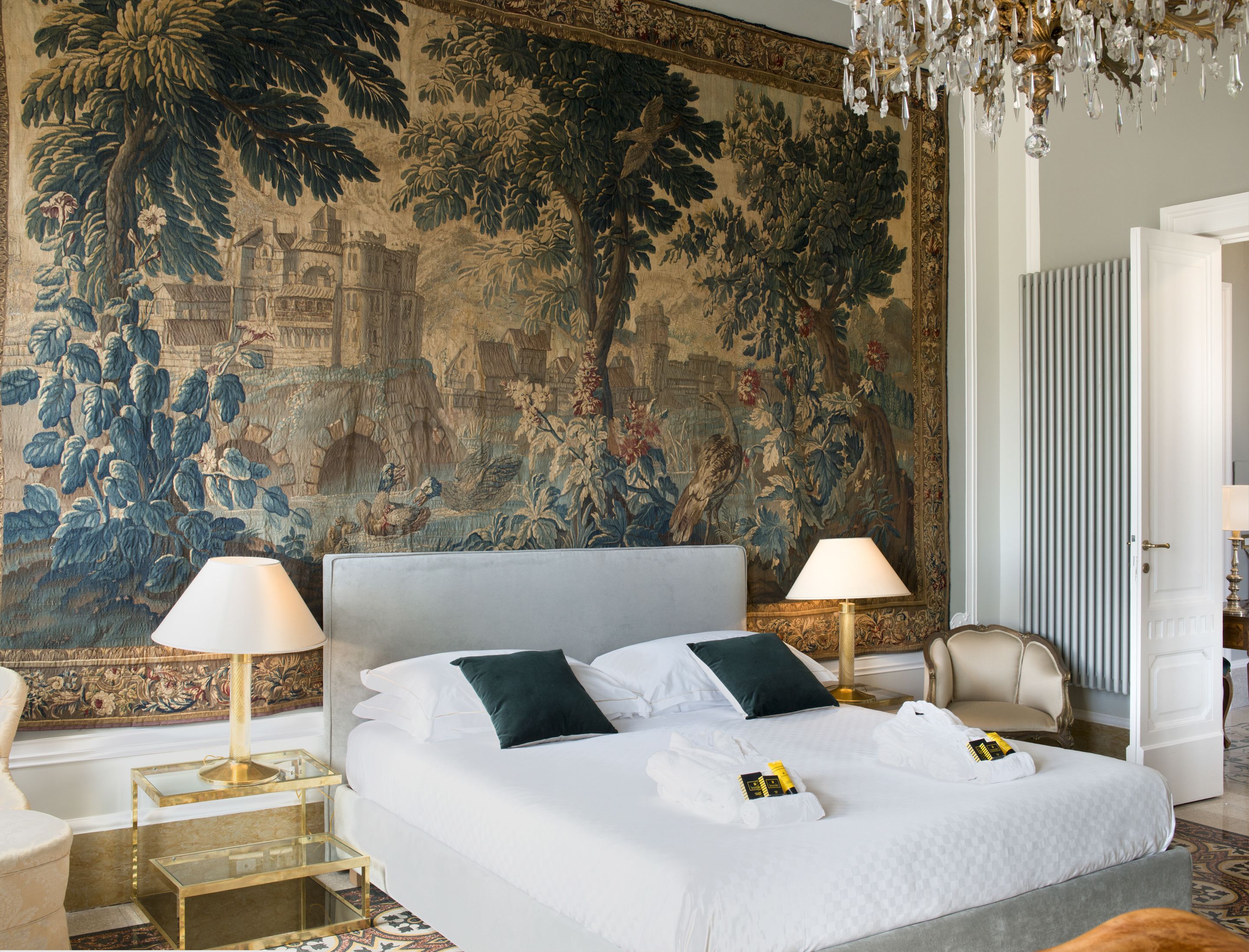 A spacious hotel room with a large bed, two bedside lamps, a chandelier, and a detailed tapestry on the wall.